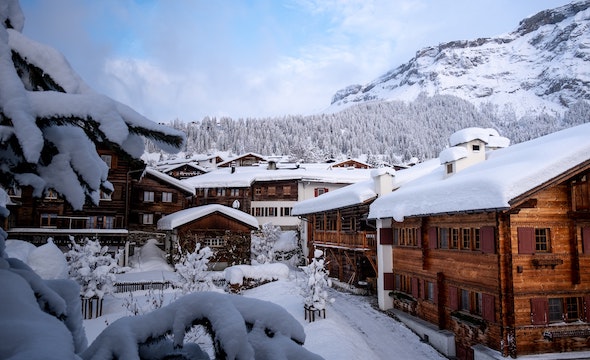 A scenic winter village scene featuring traditional wooden chalets covered in heavy snow, with snow-laden trees in the foreground and a forested mountain in the background under a partly cloudy sky.