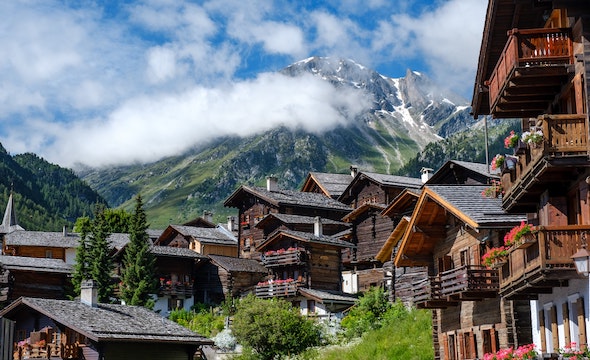 A picturesque alpine village in summer with wooden chalets adorned with flowers, nestled on a green hillside, with a cloud-covered mountain rising in the background under a blue sky.